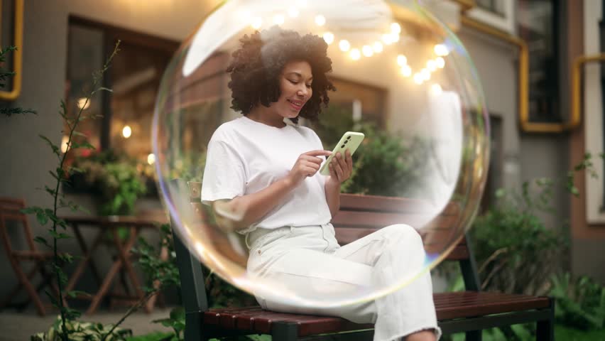 Young woman sits inside a transparent bubble while texting on her phone, representing the concept of digital or social media echo chambers