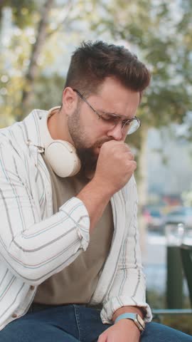 Caucasian young adult man tourist coughing while sitting on bench in city. Sick male suffers from pneumonia or inflammation of the lungs. Traveler is unwell, has a fever, a cold, a virus or the flu.