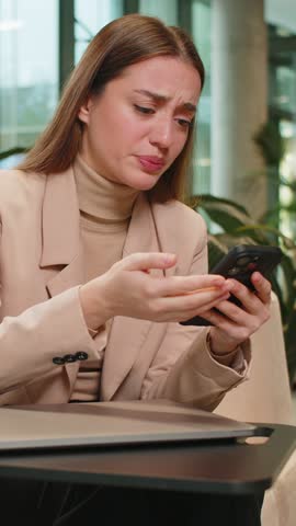 Caucasian young businesswoman girl using smartphone typing browsing, loses becoming surprised sudden lottery results, bad news, fortune loss, fail in modern office lobby. Lady sitting in hotel hall