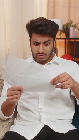 Indian man at home looking at bills with shock, throwing receipts in frustration over financial stress. Arabian guy on sofa feeling anxious, struggling with debt and mistakes in financial reports