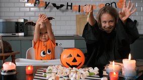 Two children dressed in Halloween costumes are sitting at a table in a decorated kitchen. They pretend to scare, surrounded by a variety of candies and spooky decor, slow motion - Powered by Shutterstock - Get 15% off with code: PIKWIZARD15