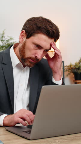 Middle-aged businessman at home office reads pay cut email on laptop and sighs after bad news sadly. Freelancer guy at table frowns thinking about salary cut and next steps for bills budget and rent.