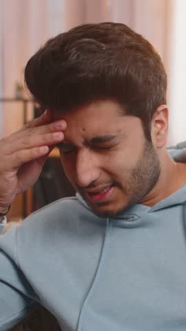 Sick Indian man rubbing temples to cure headache, suffering from tension, migraine, stress, and high blood pressure at home. Hispanic guy holds head, grimacing in pain, experiencing severe discomfort.