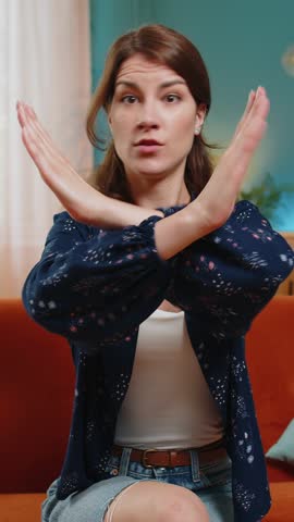 Stop gesture. Angry Caucasian woman says No hold palm folded crossed hands, warning of finish, prohibited access, declining communication, body language, danger. Girl at home room sits on sofa couch.