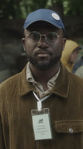 Vertical portrait of Black young male volunteer wearing blue cap, glasses and badge looking at camera while organising outdoor food kitchen for people in need