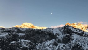 Stunning landscape view of snow-covered mountain peaks illuminated by the golden light of sunrise (alpenglow), with the moon visible in the clear blue sky. A serene and majestic alpine scene. - Powered by Shutterstock - Get 15% off with code: PIKWIZARD15
