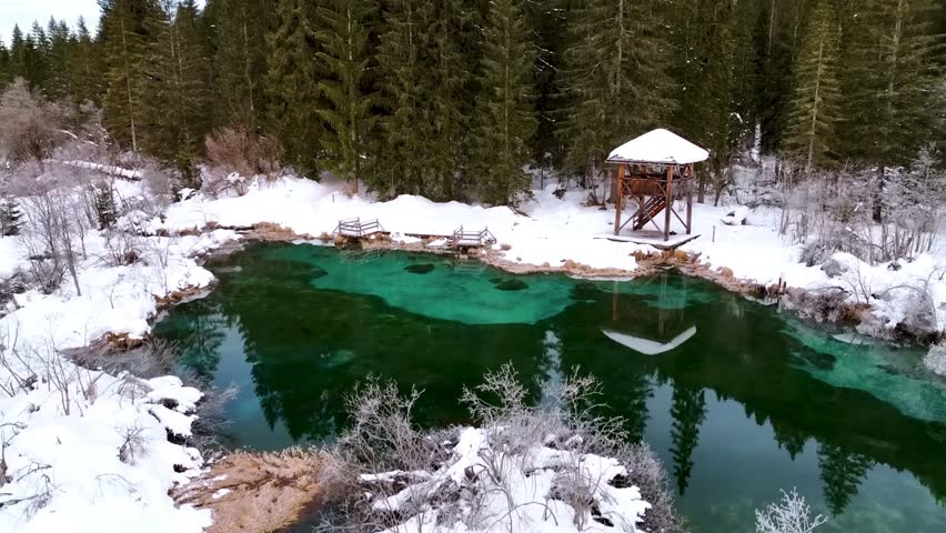 Magical aerial drone footage of a natural spring with incredibly clear turquoise and emerald water, surrounded by a snow-covered landscape and a dense pine forest. A hidden paradise in winter.