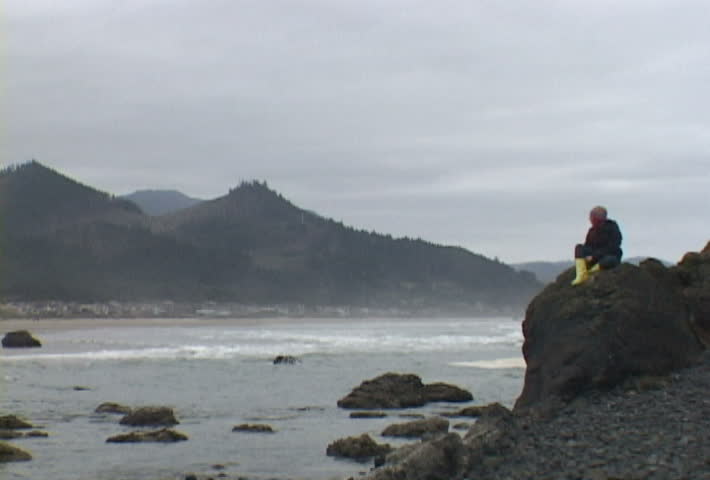 Woman at Haystack Rock in Cannon Beach, Oregon sits looking out by ocean and