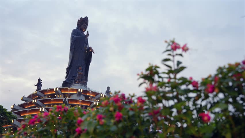 A colossal bronze statue of a Bodhisattva stands majestically on a tiered, illuminated base on Ba Den Mountain, framed by vibrant flowers in the foreground.
