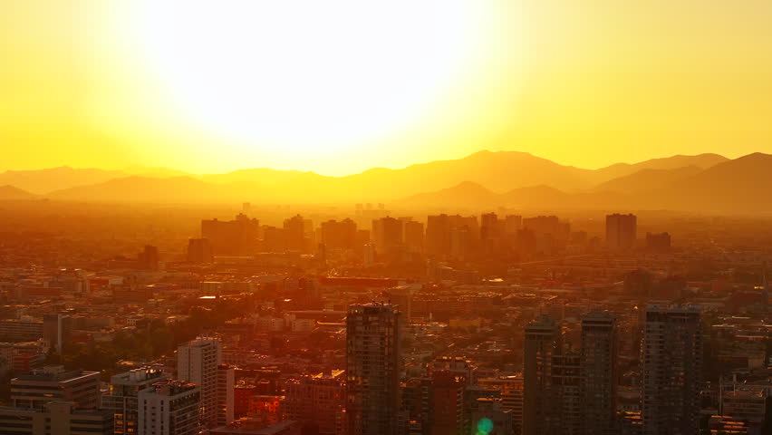Golden hour aerial shot of Santiago de Chile during a summer sunset, with the sun setting behind the Andes and tall buildings casting long shadows over the sprawling cityscape