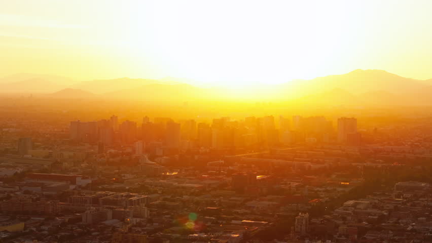 Golden hour aerial shot of Santiago de Chile during a summer sunset, with the sun setting behind the Andes and tall buildings casting long shadows over the sprawling cityscape