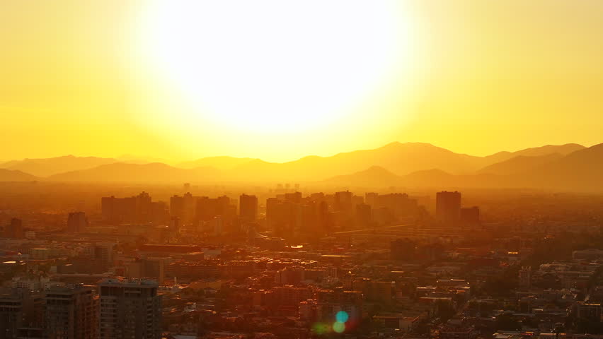 Golden hour aerial shot of Santiago de Chile during a summer sunset, with the sun setting behind the Andes and tall buildings casting long shadows over the sprawling cityscape