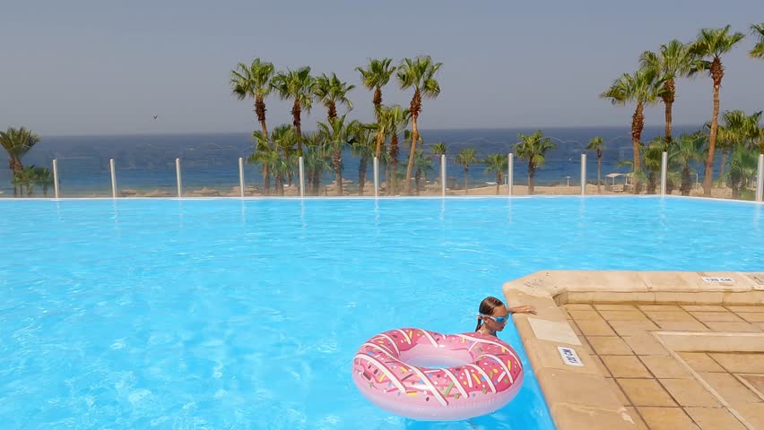 Cheerful young girl playfully entering sparkling blue swimming pool, wearing pink inflatable donut ring during sunny summer vacation day