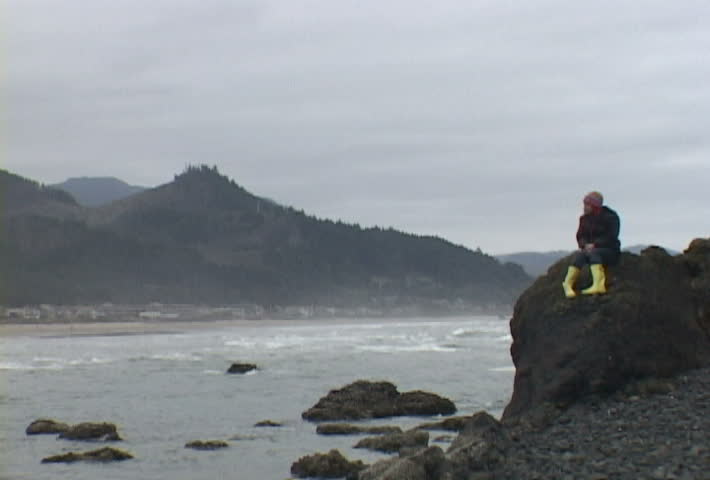 Woman at Haystack Rock in Cannon Beach, Oregon sits looking out by ocean.