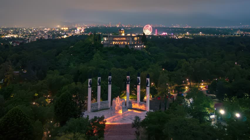 Aerial timelapse video of the Niños Héroes Monument in Chapultepec Forest, Mexico City, with the illuminated Chapultepec Castle in the background and a Ferris wheel visible in the distance.