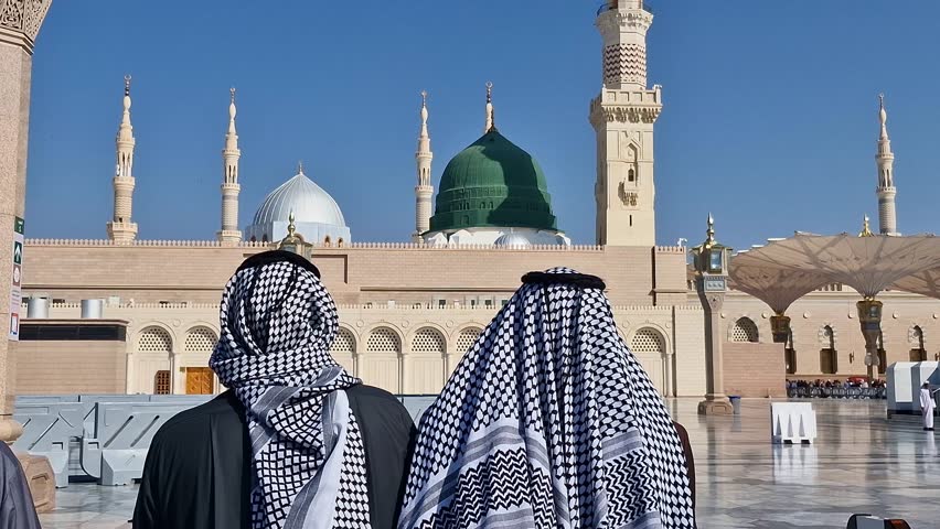 Pan up back shot view of two unidentified muslim men wearing keffiyeh look on at the green dome at the compound of Nabawi mosque in Medina, Saudi Arabia.