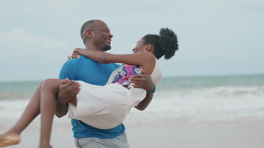 African American Couple Enjoying a Coastal Walk, Man Holding His Partner in His Arms as They Stroll Along the Beach During a Relaxing Holiday