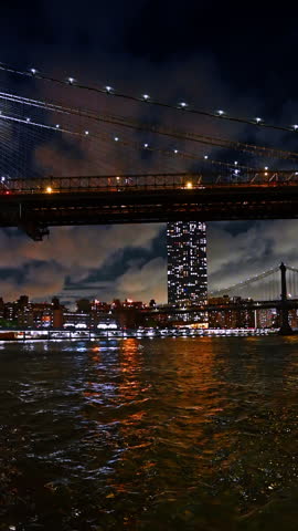Skyline of never-sleeping New York from the waterscape of the East River near the Brooklyn Bridge. Low angle view at the cloudy sky over night metropolis. Vertical video.