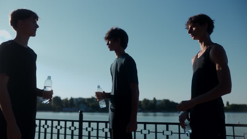 Three sporty teens pause in the park at sunset to drink water, staying hydrated after running. A healthy lifestyle shot showing energy, freshness, and togetherness.