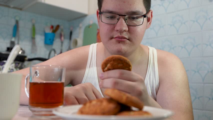 breakfast, food and people concept - happy young woman with cup of tea eating chocolate chip cookie at home over kitchen background