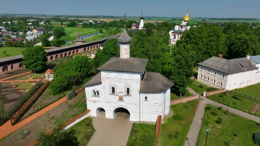 A drone's view of an ancient white-washed gatehouse church with a wooden roof, part of a large historic monastery complex in the Russian countryside on a sunny day