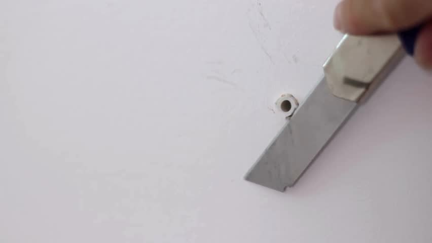 A close-up shot of a person using a utility knife to scrape away the excess plastic from a screw anchor that is secured in a white wall.