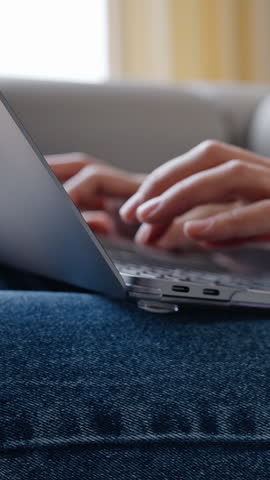 Young woman works remotely sitting on sofa in cozy apartment. Female freelancer types information to client holding laptop on lap on blurred background closeup