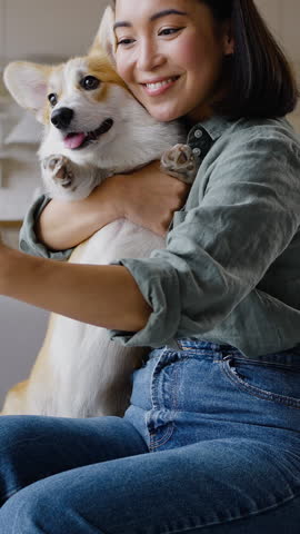 Young Asian woman takes selfie with cute dog sitting on sofa in light apartment. Smiling owner hugs Welsh corgi and kisses domestic animal lovingly low angle shot