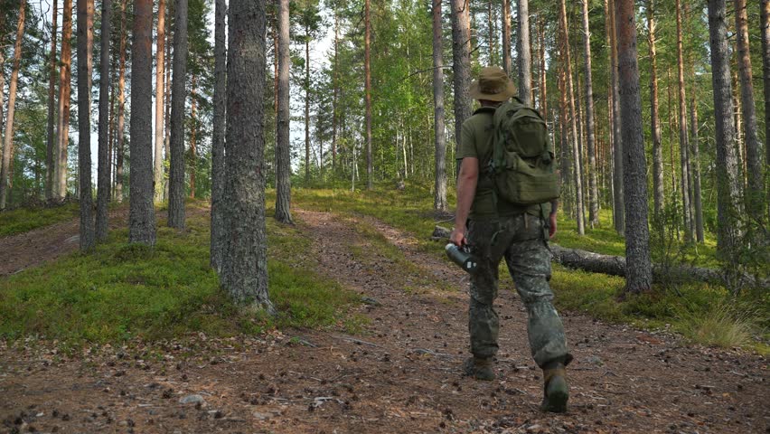 A soldier with a backpack and wearing a boonie hat is walking along a road in a coniferous forest. A man in camouflage clothing with equipment is walking along a forest trail. A ranger with a backpack