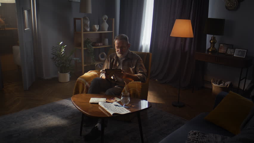 Elderly man with grey beard gazes at black-and-white photos in half-light room. Nostalgic male looks at photographs and smiles at warmth of past happiness