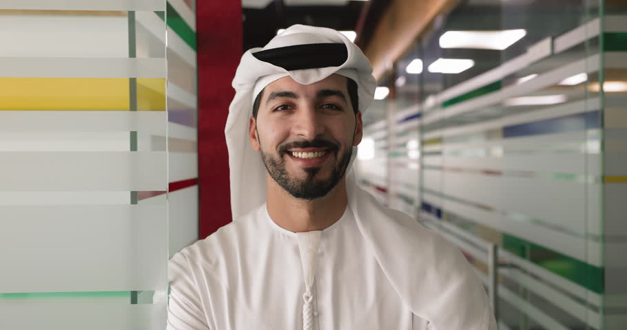 Head shot office portrait of young 30s Algerian professional businessman dressed in traditional kandura, posing in workspace hallway, exudes confidence, leadership and success in Arab business world