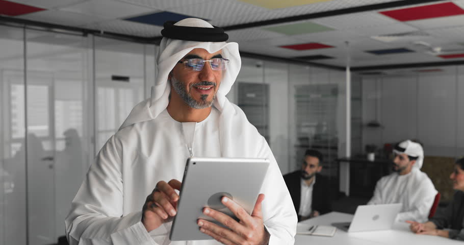 Focused middle-aged Arabic businessman in thobe stands in meeting room using digital tablet, prepare notes for negotiations, communicates with company clients or solve work-related matters remotely