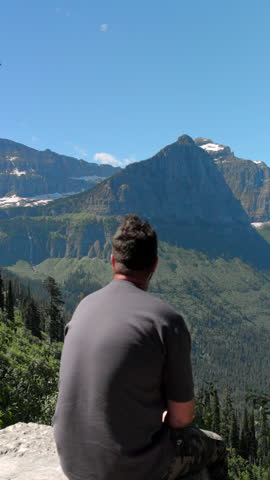Man sitting with breathtaking landscapes of Glacier National Park, Montana, featuring majestic mountains, lush greenery, and a vibrant atmosphere that captivates the soul