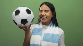 Woman holding soccer ball smiling wearing striped scarf over green background suggesting enthusiasm for sports and team spirit with a touch of youthful charm and energy. - Powered by Shutterstock - Get 15% off with code: PIKWIZARD15