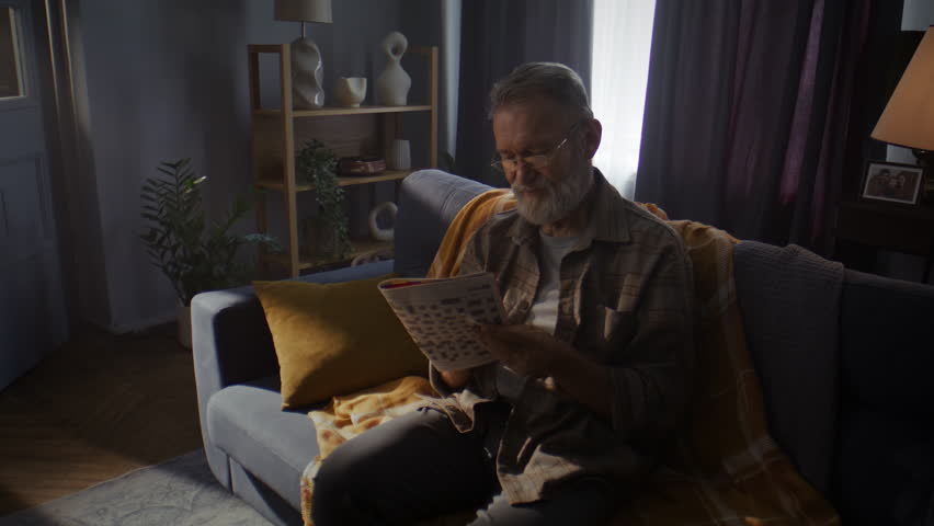 Senior man sits on comfortable sofa wrapped in blanket and reads newspaper. Pensioner with grey beard reads morning news and savors quiet atmosphere