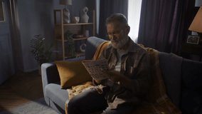 Senior man sits on comfortable sofa wrapped in blanket and reads newspaper. Pensioner with grey beard reads morning news and savors quiet atmosphere - Powered by Shutterstock - Get 15% off with code: PIKWIZARD15