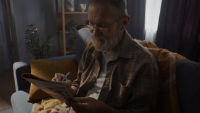 Bearded senior man fills in crossword answers with blue pen. Pensioner in glasses sits comfortably on sofa and solves clues in crossword with patience - Powered by Shutterstock - Get 15% off with code: PIKWIZARD15