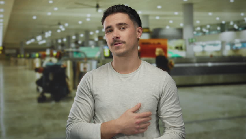Young hispanic man wearing gray shirt embraces himself in busy indoor airport terminal, highlighting themes of travel and self-reflection amidst bustling surroundings.