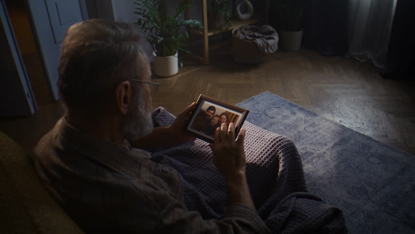 Elderly man gazes nostalgically at family photo in frame. Pensioner sits on sofa at home wrapped in blanket and recalls cherished memories from past