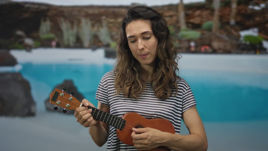 Woman gently strums ukulele by a clear blue pool in front of a hotel building under warm golden sunlight illuminating her focused expression; serenity.