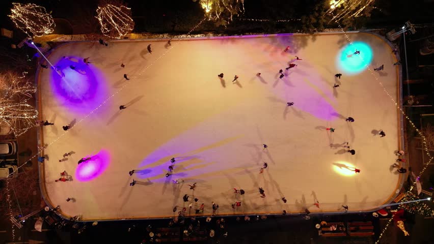Top down drone view of people skating on illuminated ice rink at night. Festive winter sports activity with colorful lights, Christmas decorations and holiday entertainment in urban outdoor setting.
