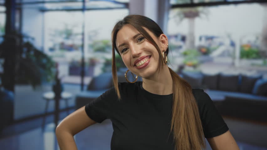 Woman smiling with teeth visible and hands on hips in a spacious building interior; confidence warmth joy positivity.