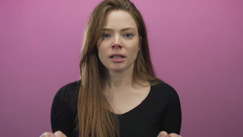 Woman clenching fists and raising open hands with a wide grin and cheerful expression in studio with pink background; joy.