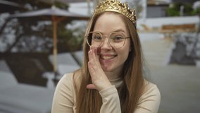 Young caucasian woman wearing golden crown presses hand to mouth leaning against building wall outdoors; secrecy joy. - Powered by Shutterstock - Get 15% off with code: PIKWIZARD15