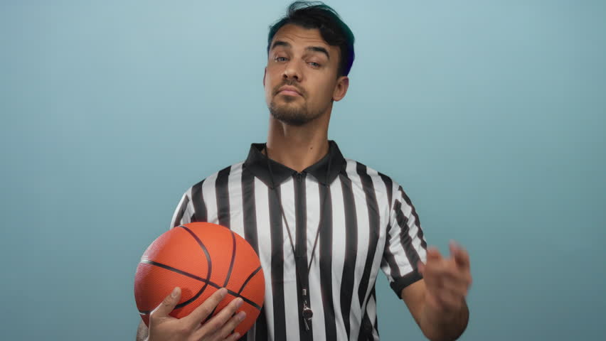 Young hispanic man holding basketball in front of isolated blue background wearing referee uniform with thoughtful expression.