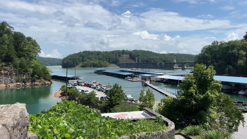 View of Norris Dam Marina at Norris Dam State Park in Tennessee with docks, parked cars, surrounding forest, and Norris Dam visible in the background.
