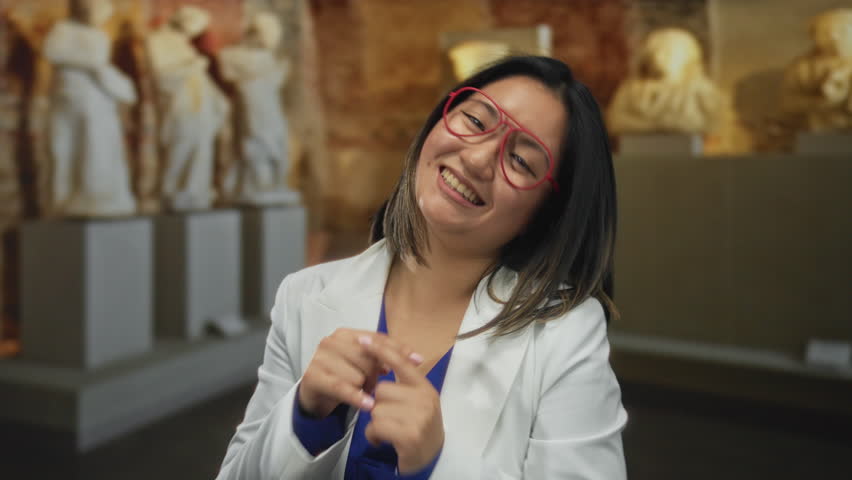 Woman wearing glasses in museum indoors playfully blowing a kiss and smiling at a camera with statues in the background.