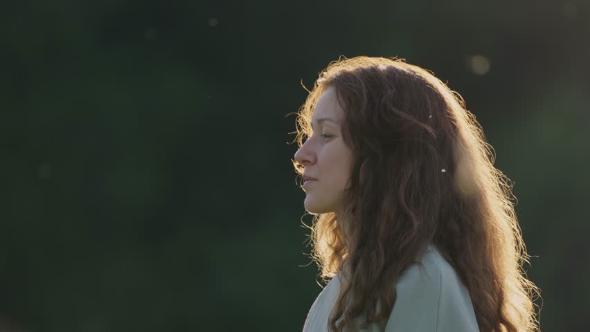 Redhead woman softly blowing dandelion seeds, golden sunset light creating dreamy atmosphere, gentle wind scattering delicate white seeds across warm landscape