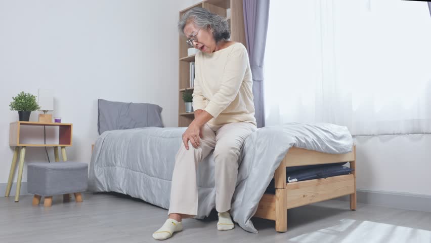 Asian Elderly woman sitting on bed holding knee pain and discomfort in bright bedroom with natural light and modern furniture creating calm and soothing atmosphere