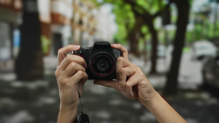 Hands holding camera on city street with blurred background, capturing an outdoor scene under dappled sunlight.